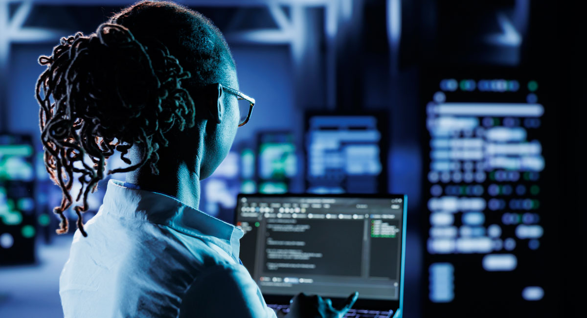 A cybersecurity professional analyzing code on a laptop while monitoring server systems in a high-tech data center.